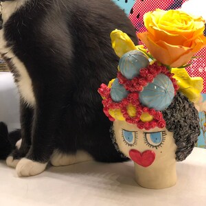 May include: A ceramic vase shaped like a woman's head with a yellow, blue, and red floral crown. The vase has a red heart-shaped nose and blue eyes. A yellow rose is in the vase.