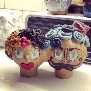 May include: A pair of ceramic figurines with painted faces. The figurine on the left has brown hair and orange and red flowers. The figurine on the right has blue and white swirls and a black mustache.