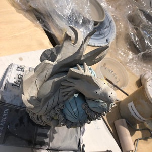 May include: A ceramic sculpture of a dragon holding a blue and white sphere. The dragon is made of gray clay and has a textured surface. The sphere is decorated with swirls and dots.
