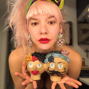 May include: A person with pink hair and a colorful headscarf is holding two ceramic figurines. The figurines are painted with colorful details and have exaggerated features. The figurine on the left is a woman with orange hair and blue eyes. The figurine on the right is a man with blue hair and a mustache.