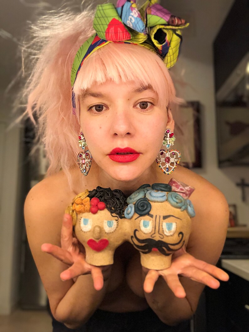 May include: A person with pink hair and a colorful headscarf is holding two ceramic figurines. The figurines are painted with colorful details and have exaggerated features. The figurine on the left is a woman with orange hair and blue eyes. The figurine on the right is a man with blue hair and a mustache.