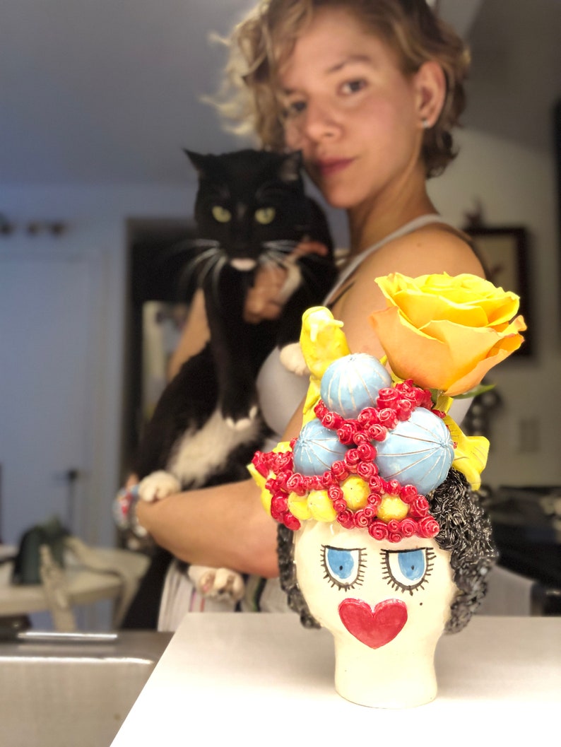 May include: A ceramic planter shaped like a woman's head with a colorful floral arrangement. The planter has a red heart, blue eyes, and a yellow face. The flowers are yellow, red, and blue.