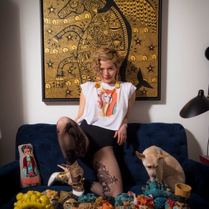 May include: A woman wearing a white t-shirt with a colorful graphic print, black shorts, and fishnet stockings poses on a blue couch. She is wearing gold earrings and has curly blonde hair. A small dog is sitting next to her. A table in front of the couch is decorated with colorful ceramic figurines with human faces.