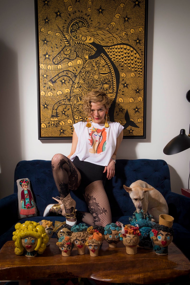 May include: A woman wearing a white t-shirt with a colorful graphic print, black shorts, and fishnet stockings poses on a blue couch. She is wearing gold earrings and has curly blonde hair. A small dog is sitting next to her. A table in front of the couch is decorated with colorful ceramic figurines with human faces.