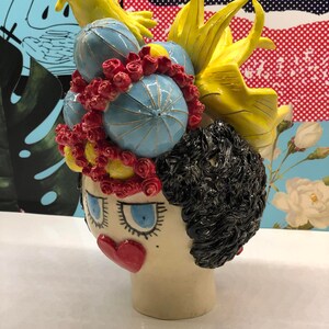 May include: A ceramic sculpture of a woman's head with a whimsical hairstyle. The sculpture is painted in bright colors, including yellow, blue, red, and black. The woman has large blue eyes and a red heart-shaped mouth.