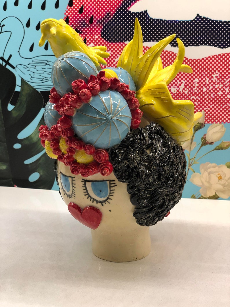 May include: A ceramic sculpture of a woman's head with a whimsical hairstyle. The sculpture is painted in bright colors, including yellow, blue, red, and black. The woman has large blue eyes and a red heart-shaped mouth.