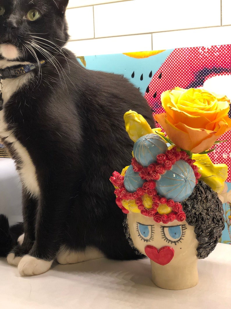 May include: A ceramic vase shaped like a woman's head with a yellow, blue, and red floral crown. The vase has a red heart-shaped nose and blue eyes. A yellow rose is in the vase.