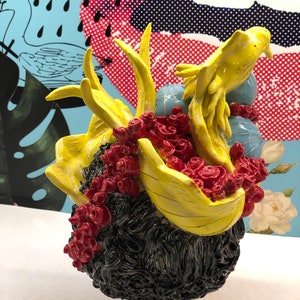 May include: A ceramic sculpture of a person's head with black hair, red flowers, and a yellow bird perched on top. The sculpture is on a white pedestal.