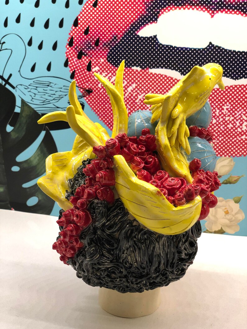 May include: A ceramic sculpture of a person's head with black hair, red flowers, and a yellow bird perched on top. The sculpture is on a white pedestal.