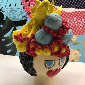 May include: A ceramic sculpture of a woman's head with a whimsical crown of yellow, red, and blue flowers. The crown features a yellow bird and blue sea urchins.