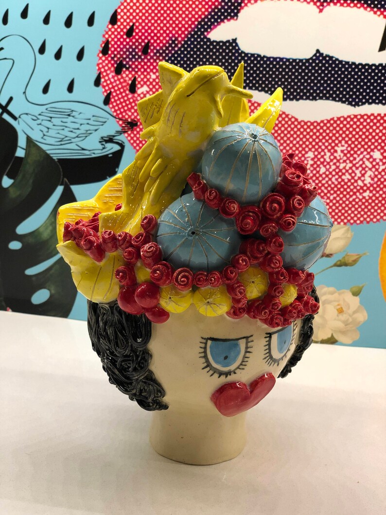 May include: A ceramic sculpture of a woman's head with a whimsical crown of yellow, red, and blue flowers. The crown features a yellow bird and blue sea urchins.