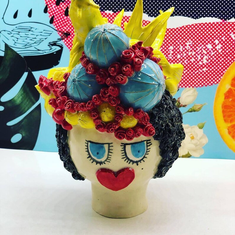 May include: A ceramic figurine of a woman's head with a whimsical hairstyle. The hair is styled with yellow, blue, and red flowers and leaves. The woman has large blue eyes, a red heart-shaped nose, and a white face.