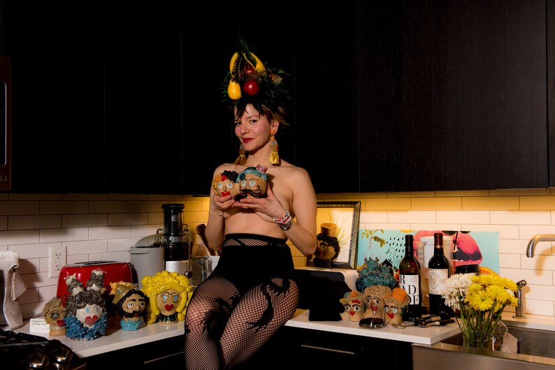 May include: A woman wearing a black skirt and fishnet stockings holds three ceramic figurines with painted faces. She is wearing a crown of fruit and vegetables on her head. The figurines are sitting on a counter with other figurines, wine bottles, and flowers.