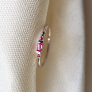 Op de afbeelding: Een delicate zilveren ring met een rechthoekige roze edelsteen en kleinere roze edelstenen aan beide kanten.
