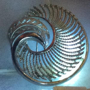 May include: A silver-toned brooch with a swirling, abstract design. The brooch is made of metal and has a delicate, intricate pattern.