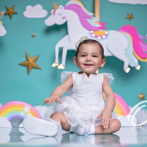 Puede incluir: Una niña pequeña con un vestido blanco se sienta frente a un fondo turquesa con decoraciones de unicornio y arcoíris. El texto "One" está escrito en blanco sobre una nube.