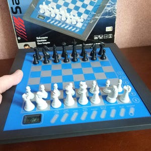 May include: A chess computer with a blue and gray checkered board and black and white chess pieces. The board has a digital display and is framed in black. The box in the background shows a similar board.