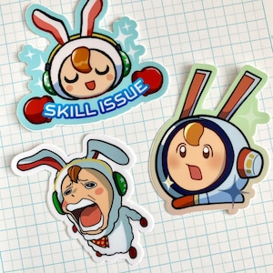 May include: Three cartoon character stickers with bunny ears and a white background. The first sticker has a character with a red and white bunny ears, green headphones, and the text "SKILL ISSUE". The second sticker has a character with brown bunny ears and a blue and white space suit. The third sticker has a character with blue bunny ears, green headphones, and a white sheep costume.
