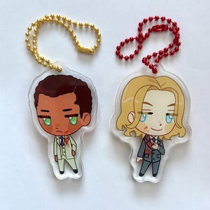 Pode incluir: Dois chaveiros de acrílico. Um apresenta um homem de desenho animado com cabelo castanho em um terno branco, o outro um homem de desenho animado com cabelo loiro em um terno cinza. Ambos têm olhos verdes e são presos a uma corrente dourada e vermelha.