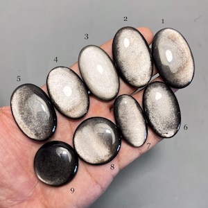 Op de afbeelding: Een verzameling van negen gepolijste, ovale en ronde edelstenen in tinten zwart, zilver en goud. De stenen variëren in grootte en vorm, met een reflecterende, metallic glans. De edelstenen worden op een neutrale achtergrond gepresenteerd.