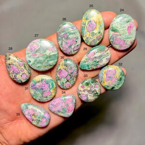 May include: A collection of polished gemstones in various shapes, including oval, round, and teardrop. The stones feature a mix of pink, green, and cream colors, with a marbled appearance. The gemstones are held in a hand.