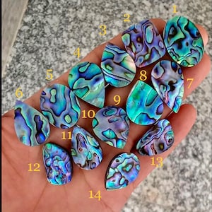 May include: A collection of fourteen iridescent abalone shell cabochons in various shapes, including teardrop, oval, and square. The shells display a vibrant mix of blues, greens, and purples, with swirling patterns. The cabochons are numbered 1 through 14.