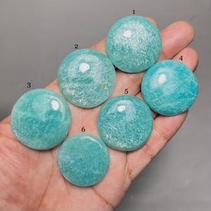 Puede incluir: Se muestran seis piedras preciosas redondas y pulidas en tonos turquesa y verde azulado. Las piedras tienen una superficie lisa y brillante con sutiles vetas. Las piedras preciosas se sostienen en una mano.