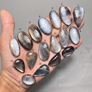 Op de afbeelding: Een verzameling van zeventien gepolijste edelstenen in verschillende vormen, waaronder ovaal, druppelvormig en driehoekig. De stenen vertonen een reeks kleuren, zoals goud, bruin en blauw, met reflecterende oppervlakken. De edelstenen zijn genummerd van 1 tot 17.