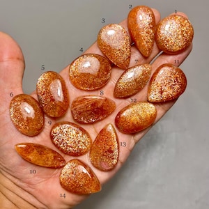 Op de afbeelding: Een verzameling van veertien gepolijste zonnesteen edelstenen in verschillende vormen, waaronder ovaal, druppelvormig en rond. De stenen vertonen een warme oranje tint met sprankelende insluitsels, waardoor een glinsterend effect ontstaat. De edelstenen worden in een hand gehouden.