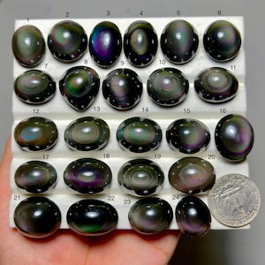 May include: A collection of twenty-four iridescent obsidian gemstones in various oval and round shapes, displayed on a white tray. The stones exhibit a range of colors, including green, purple, and gold, with a quarter coin for size comparison.