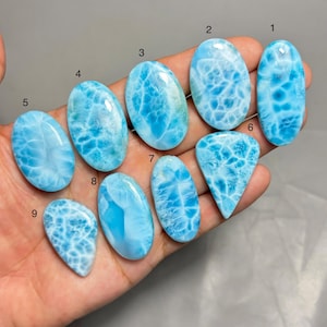 Könnte beinhalten: Eine Sammlung von neun polierten Larimar-Edelsteinen in verschiedenen Formen, darunter oval und tropfenförmig. Die Steine zeigen eine Reihe von hellblauen Farbtönen mit weißen, wolkenartigen Mustern. Die Edelsteine werden in einer Hand gehalten.
