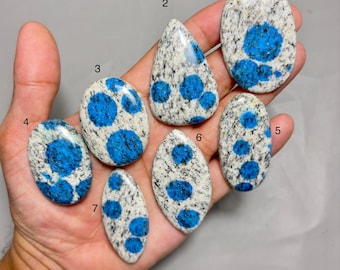 May include: A collection of seven polished k2 jasper stones. Each stone features a white and gray speckled background with vibrant blue orb-like patterns. The stones vary in shape, including oval and teardrop designs.