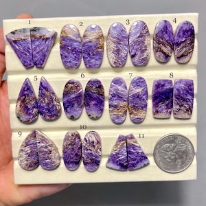 May include: A collection of eleven polished charoite gemstones in various shapes, including ovals, teardrops, and trapezoids. The stones display a mix of deep purple, brown, and white swirling patterns. A US quarter is included for size reference.