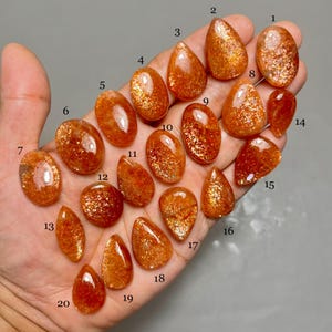 May include: A hand displays a collection of polished sunstone gemstones in various shapes, including oval, teardrop, and pear. The stones exhibit a warm orange hue with shimmering inclusions, creating a sparkling effect. The gemstones are numbered for identification.