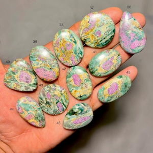 Può includere: Una collezione di pietre preziose lucide di varie forme, tra cui ovali, a goccia e rotonde. Le pietre presentano un mix di tonalità verdi, rosa e gialle, con un aspetto marmorizzato. Le pietre preziose sono tenute in una mano, mostrando i loro motivi e colori unici.