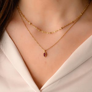 Op de afbeelding: Een goudkleurige ketting met twee lagen en een kleine, ovaalvormige, rode edelsteenhanger. De bovenste laag heeft kleine gouden kralen, terwijl de onderste laag de hanger vasthoudt. De ketting wordt getoond tegen een lichte achtergrond.