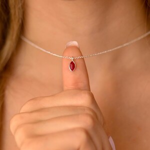 May include: A silver necklace with a small, red, marquise-cut gemstone pendant. The pendant hangs from a fine chain. The necklace is displayed against a blurred background.
