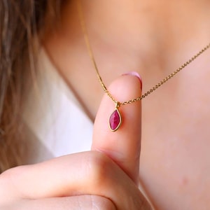May include: A gold-colored necklace featuring a small, teardrop-shaped pendant with a vibrant red gemstone. The pendant is suspended from a delicate chain. The necklace is displayed on a person's finger.