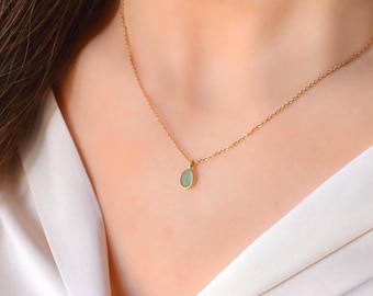 May include: A delicate gold-colored necklace featuring a small, oval-shaped pendant with a pale green gemstone. The pendant hangs from a fine chain, resting against a person's skin, and is set against a white background.