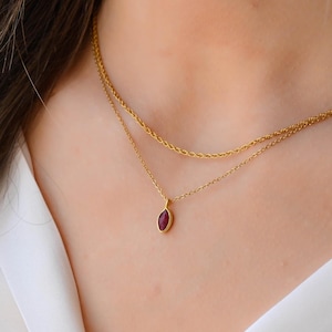 Peut inclure: Un collier doré à deux chaînes. La chaîne inférieure porte un pendentif avec une pierre de couleur marron en forme d'ovale. Le collier est porté sur une chemise blanche.