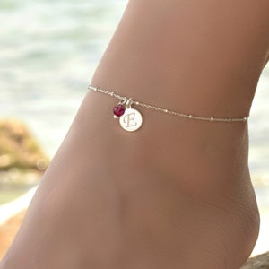 Puede incluir: Tobillera de plata con un pequeño dije redondo grabado con la letra "E". Una pequeña piedra preciosa de color rojo oscuro cuelga del dije.