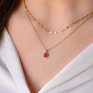 Peut inclure: Un collier en chaîne dorée avec un pendentif en forme de larme orné d'une pierre précieuse rouge. Le collier est superposé à une chaîne dorée délicate.