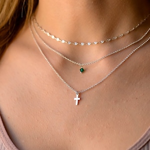 May include: Three layered necklaces. The top necklace is a delicate chain with small, oval-shaped embellishments. The middle necklace features a small, green gemstone pendant. The bottom necklace has a small cross pendant.