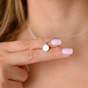 May include: A silver necklace with a small, round pendant. The pendant features the letter "C" in a simple, elegant font. The necklace is displayed against a blurred background.
