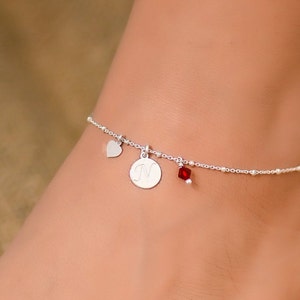 Puede incluir: Tobillera de plata con un pequeño dije de corazón, un dije redondo con la letra "N" y un dije de cristal rojo.