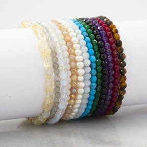 May include: A stack of colourful beaded bracelets in various shades of yellow, white, blue, green, black, purple, and brown. The bracelets are made of natural stones and are stacked on a white surface.