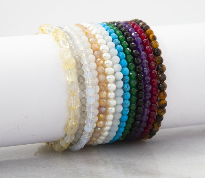 May include: A stack of colourful beaded bracelets in various shades of yellow, white, blue, green, black, purple, and brown. The bracelets are made of natural stones and are stacked on a white surface.
