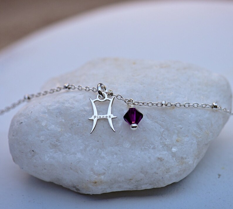 Puede incluir: Un brazalete de cadena de plata con un dije de Piscis y una cuenta de cristal morado. El dije est&aacute; grabado con la palabra "Pisces".