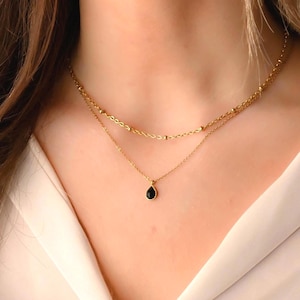 May include: A gold chain necklace with a small black teardrop pendant. The necklace is layered with a second gold chain with small beads.