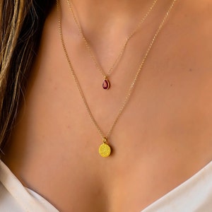 May include: Two gold necklaces layered together. The top necklace features a small, teardrop-shaped pendant with a red gemstone. The bottom necklace has a textured, circular pendant. Both necklaces are delicate and elegant.